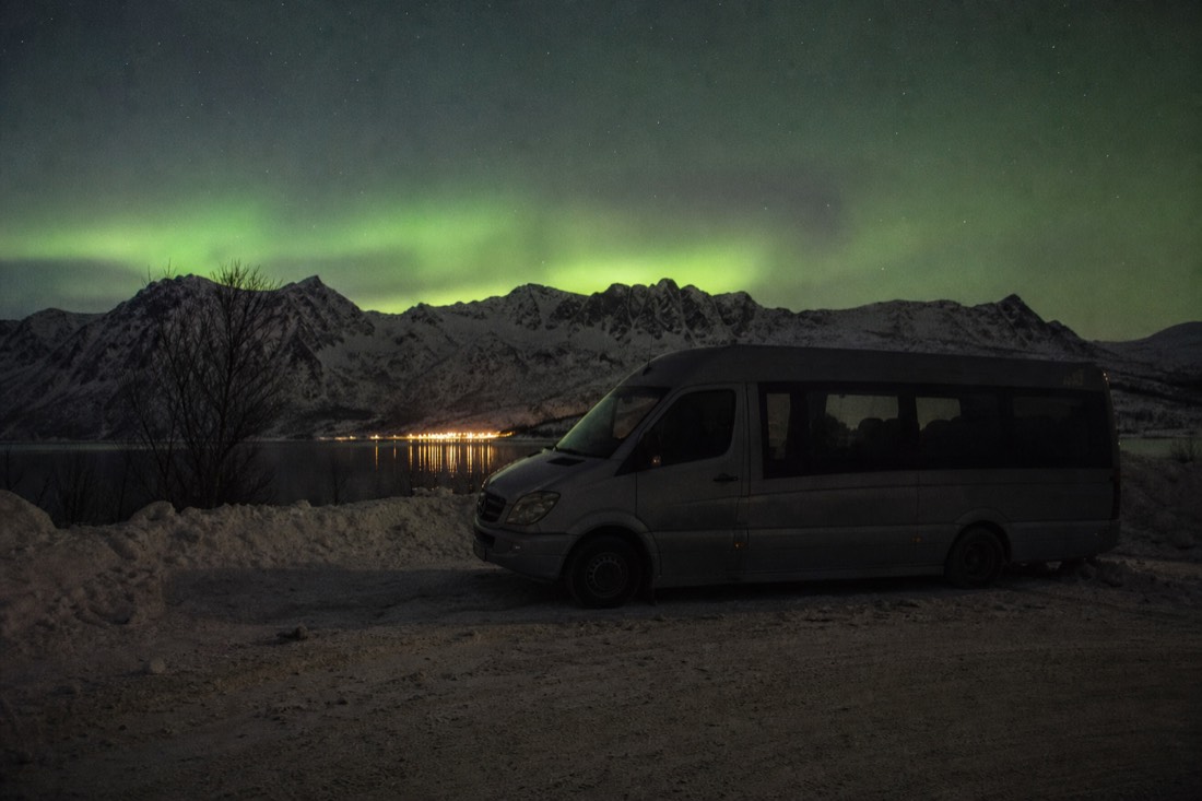 Minibus parked on a snowy roadside at night with headlights off and a faint green glow above the ridge