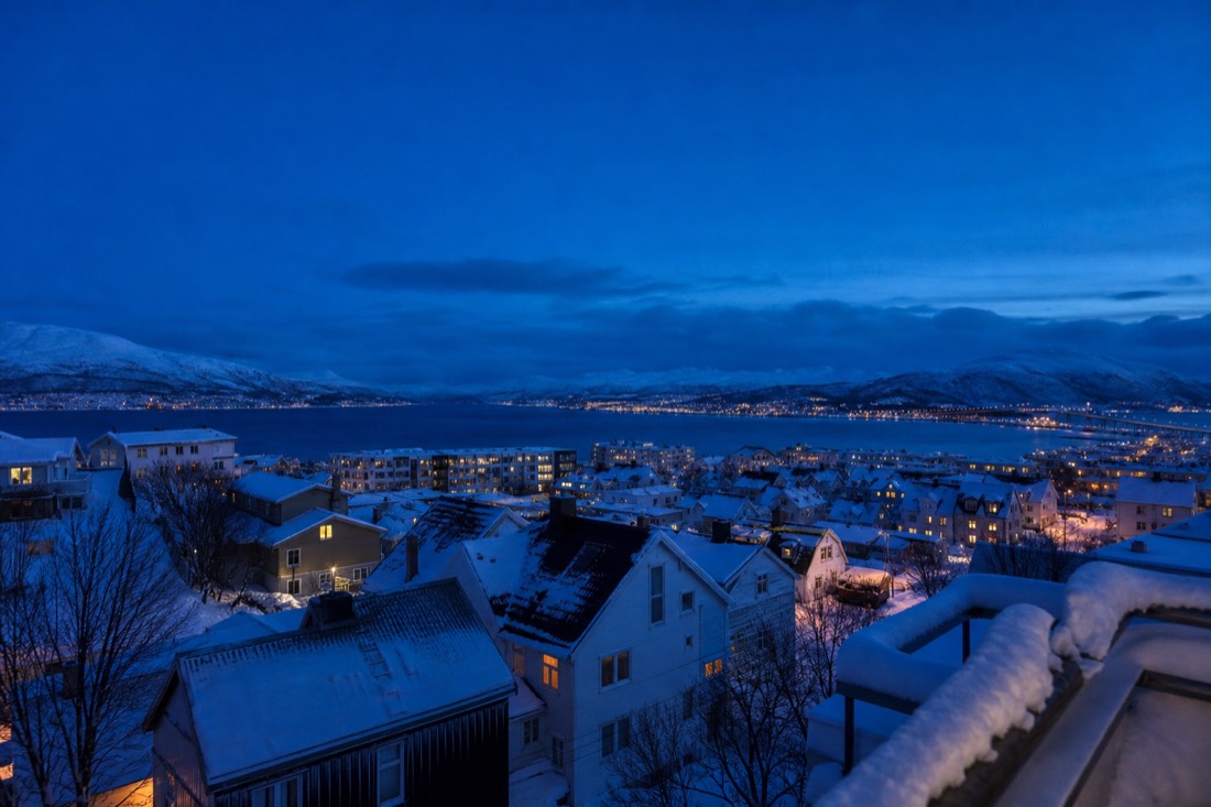 Blue midday twilight over Tromso rooftops in December with warm lit windows and snow on the railings