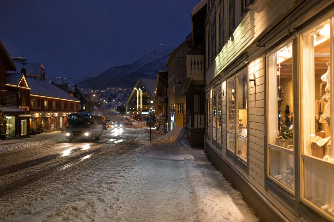 Snowy Tromso street at night with lit shop windows and a minibus waiting at the kerb