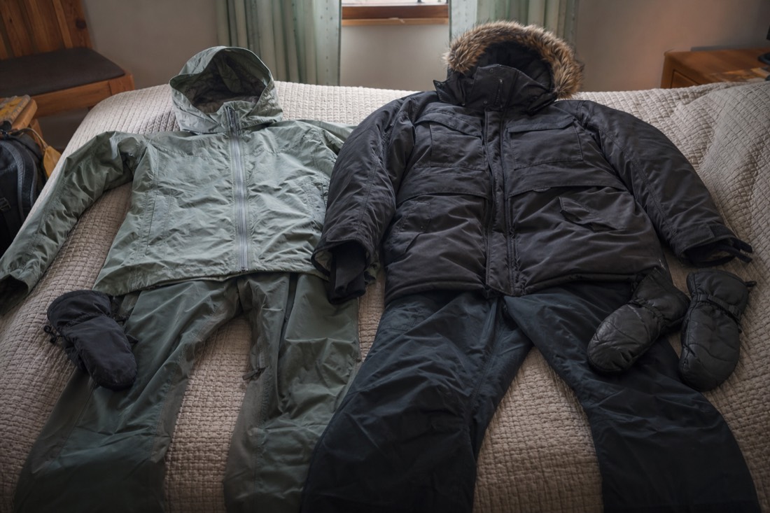 Two sets of outdoor clothing laid side by side on a bed, a light rain shell and a heavy insulated parka