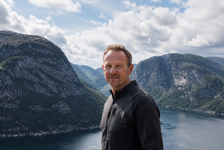 Lars Hansen standing above a western Norwegian fjord, steep green mountainsides and deep blue water behind him
